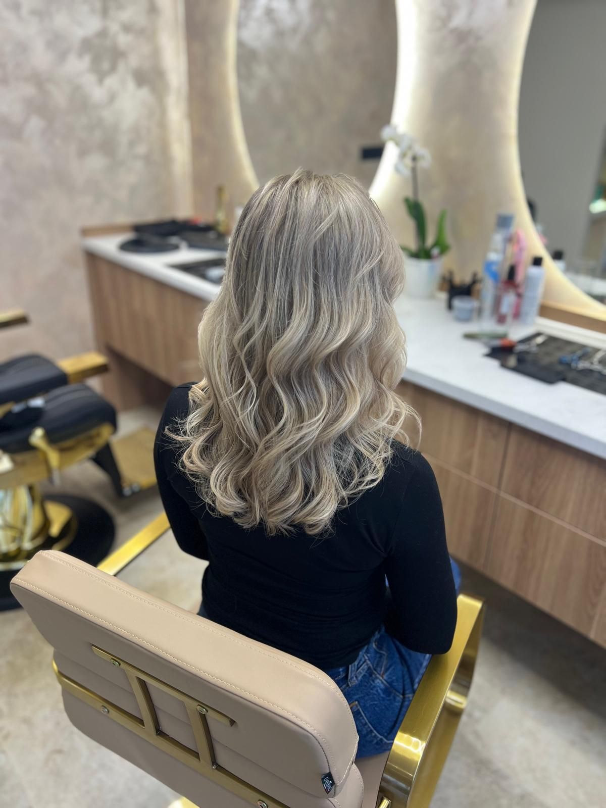 Klientka s dlhými blond vlnami v kresle pred oválnym zrkadlom salóna Hair Essence